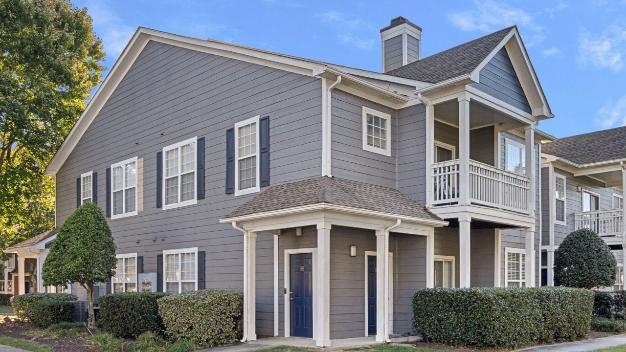 Two-story gray apartment building with white trim, balconies, and bushes along the sidewalk on a sunny day.