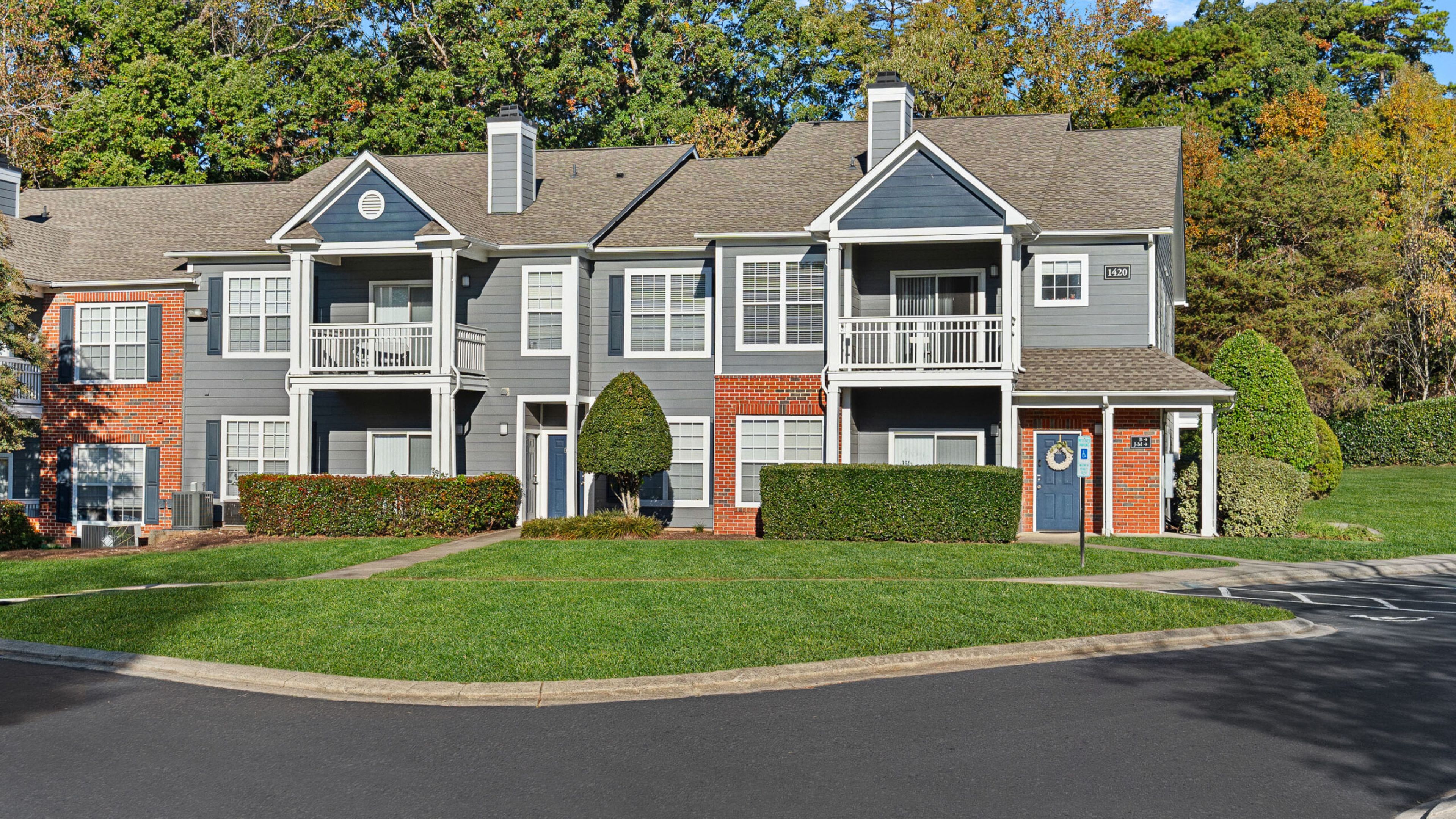 Two-story apartment building with gray siding, brick accents, balconies, and trimmed bushes in front.