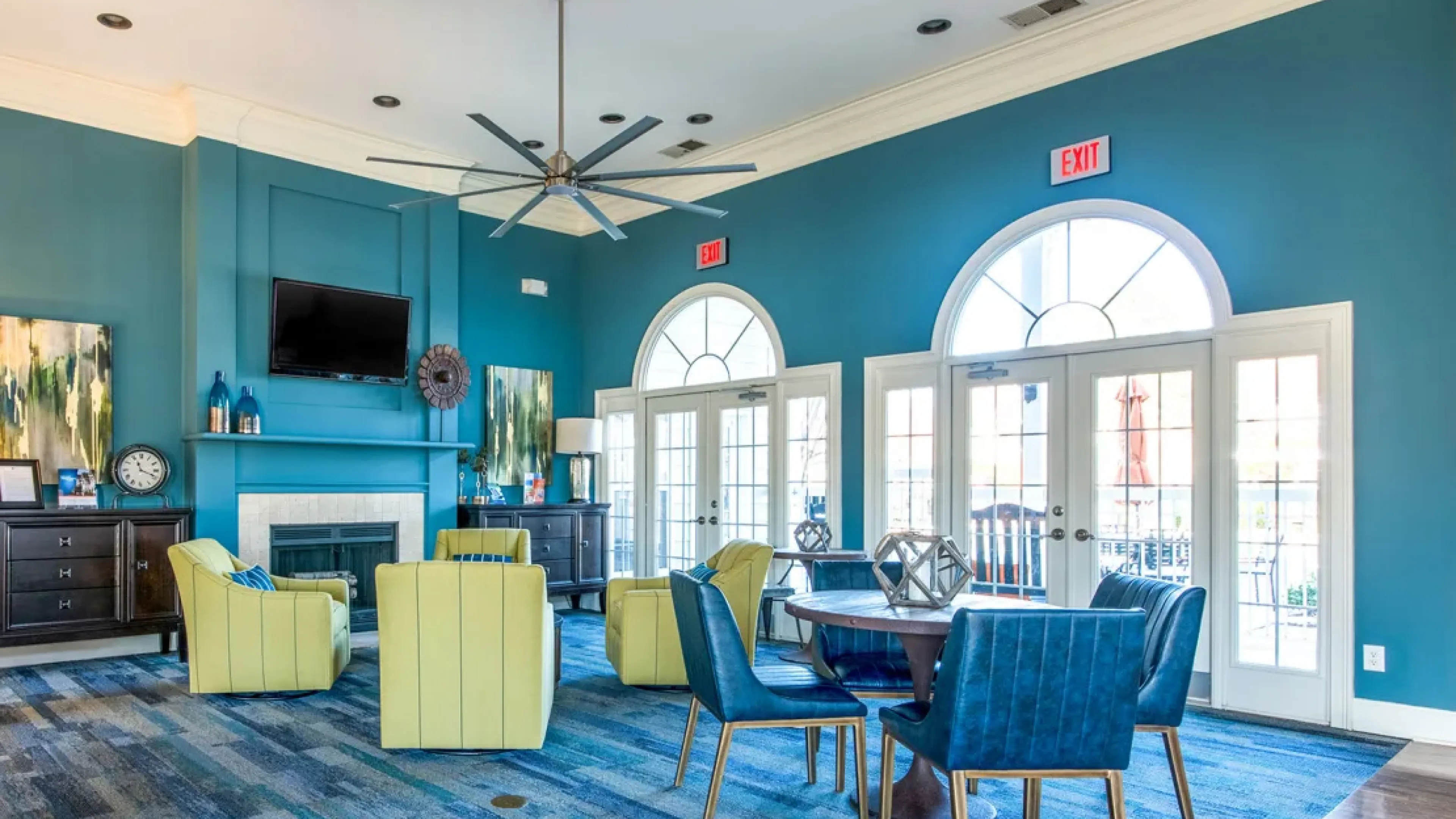 Modern lounge with blue walls, yellow chairs, blue chairs, a fireplace, TV, and large bright windows.
