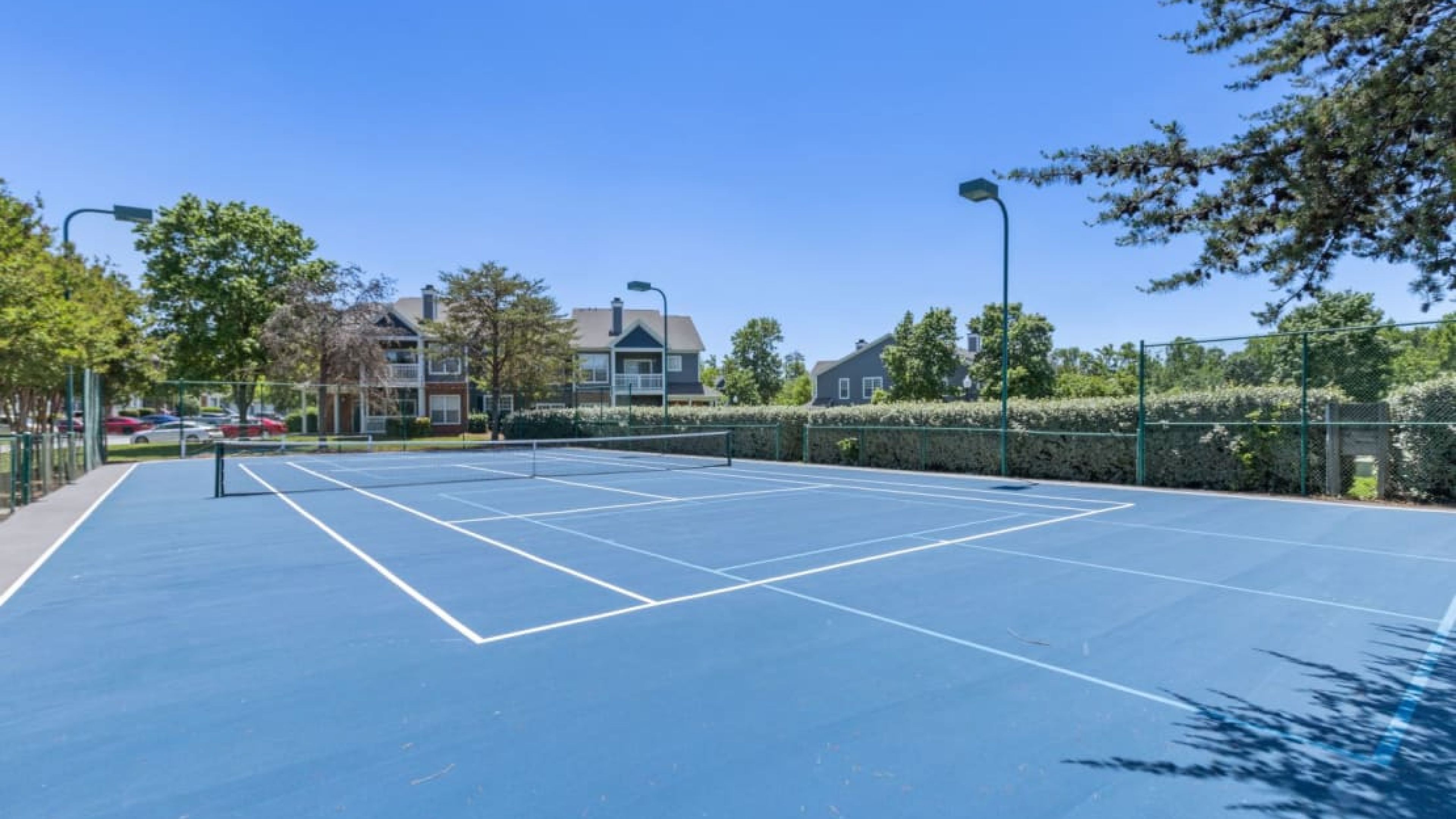 Blue outdoor tennis court with white lines, surrounded by trees and residential buildings under a clear sky.